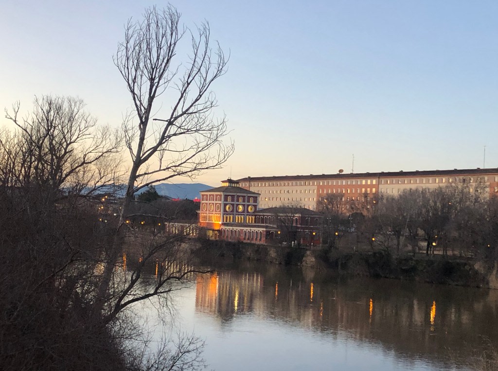 An image of the river with a colorful building at sunset, surrounded by trees and mountains in the background.