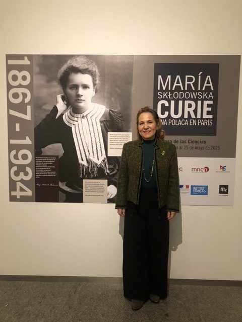A woman posing in front of an informational panel about Maria Sklodowska-Curie at an exhibition, with photographs and explanatory texts about her life and work.