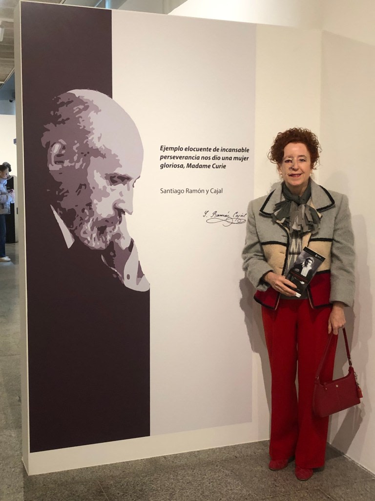 A person standing next to a panel presenting a quote from Santiago Ramon y Cajal about Madame Curie, with an illustration of Cajal in the background. The person holds a brochure and wears a light jacket with red trousers.