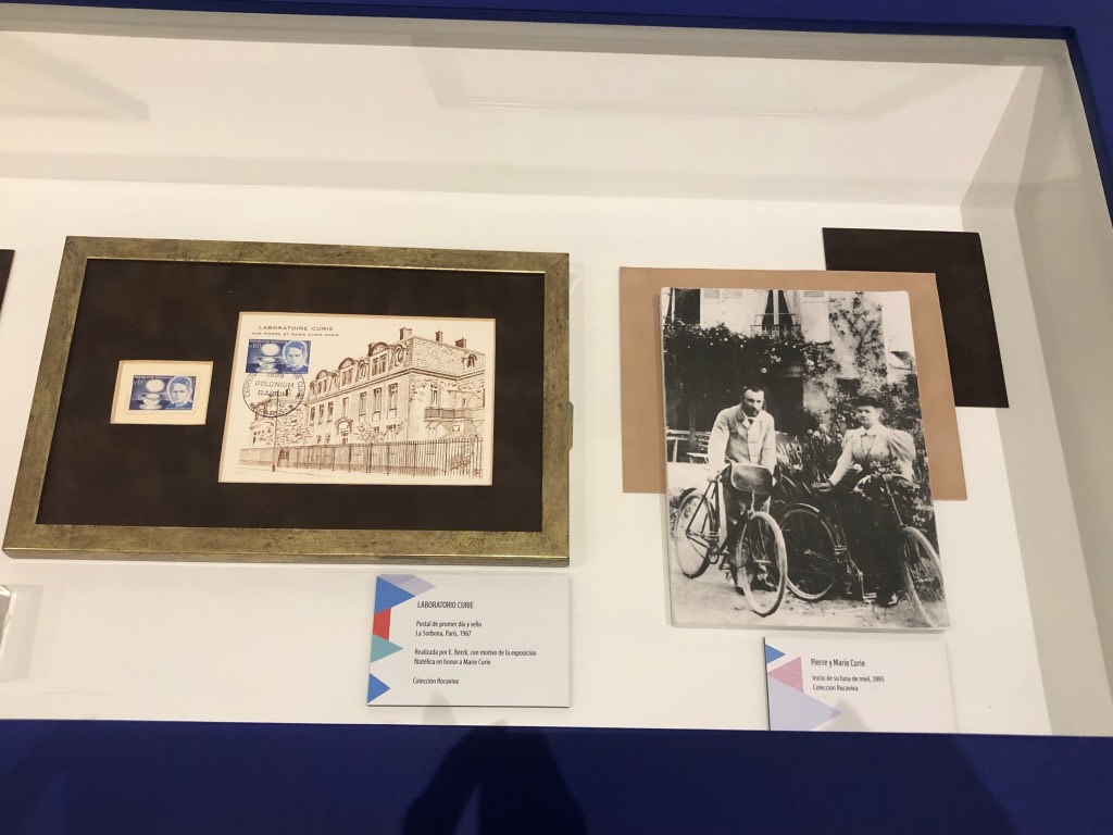 Display of a letter and a commemorative stamp from the Curie laboratory, alongside a photograph of Pierre and Marie Curie on a bicycle, in a display case at the Maria Sklodowska-Curie exhibition.