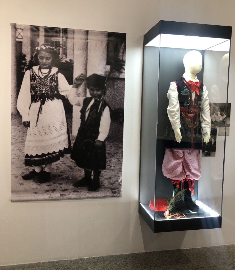A black and white photograph of two children in traditional costumes, one of them holding a branch, next to a display case exhibiting a traditional Polish costume adorned with medals and cultural accessories.