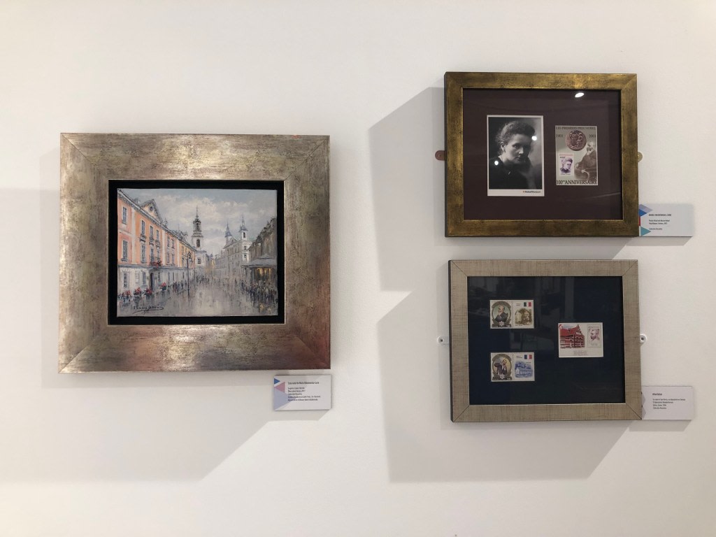 A painting of an urban scene, framed, next to two frames with documents and philatelic stamps related to Marie Curie, displayed on a white wall.