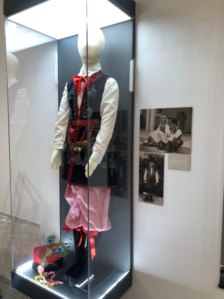 Traditional Polish costume exhibited in a display case, with colorful adornments and a hairstyle next to a photograph of children dressed traditionally.