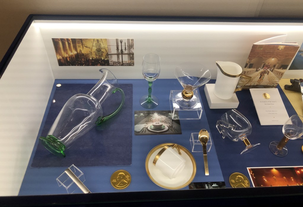 Display of objects and memorabilia from the Nobel ceremony, including a glass jug, cups, plates, and a gold medal, in a display case at the Maria Sklodowska-Curie exhibition.