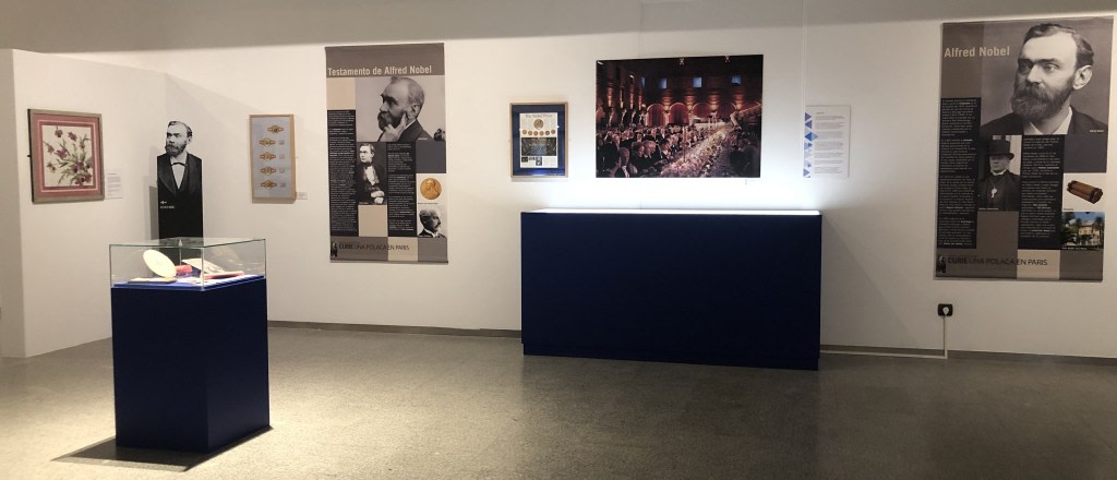 Exhibition dedicated to Alfred Nobel, with informational panels and objects related to his life and legacy, including a reproduction of his will and a display case with antique pieces. A museum setting with appropriate lighting is observed.