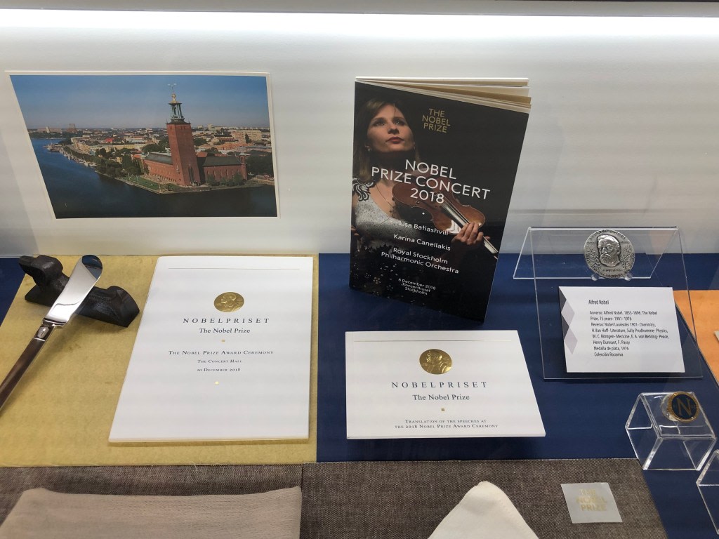 Display of objects related to the Nobel Prize, including pamphlets, a medal, and a program from the 2018 award concert, with a backdrop of the city of Stockholm.