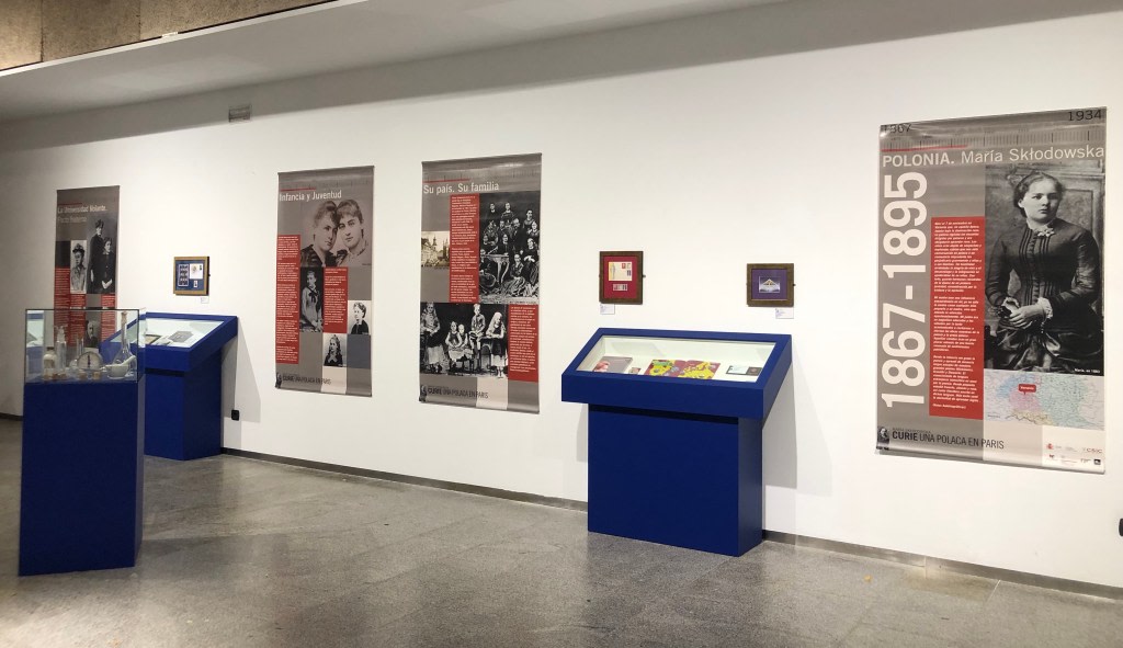 Exhibition dedicated to the life and work of Maria Sklodowska-Curie, with informational panels and display cases showing objects related to her history, in an educational setting.