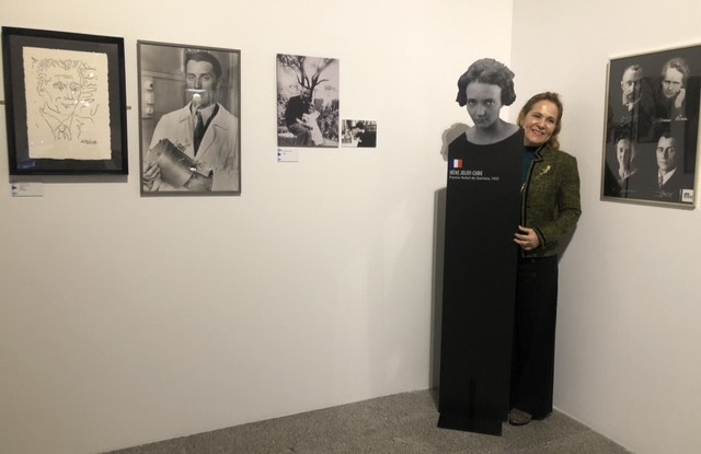 Person posing next to a figure of Maria Sklodowska-Curie at an exhibition, with various photographs and a portrait on the wall.