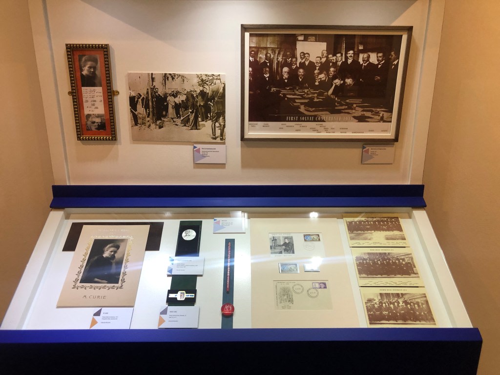 Exhibition display case showing a collage of documents related to Maria Sklodowska-Curie, including photos, stamps, and a framed portrait of the scientist.