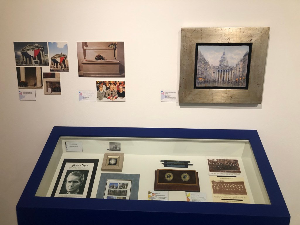 Exhibition about Maria Sklodowska-Curie, with photographs and framed documents, next to a display case exhibiting objects related to her life and achievements.