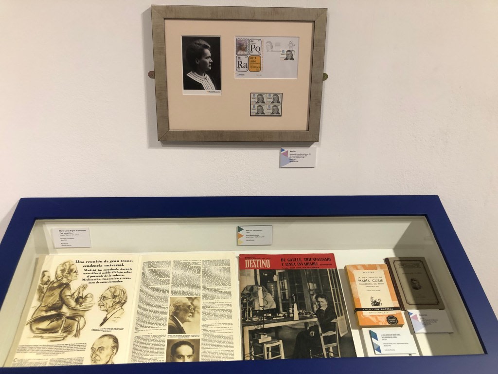 Display of documents and photographs related to Maria Sklodowska-Curie, including a portrait, stamps, and scientific journals, in a display case at the exhibition about her life and work.