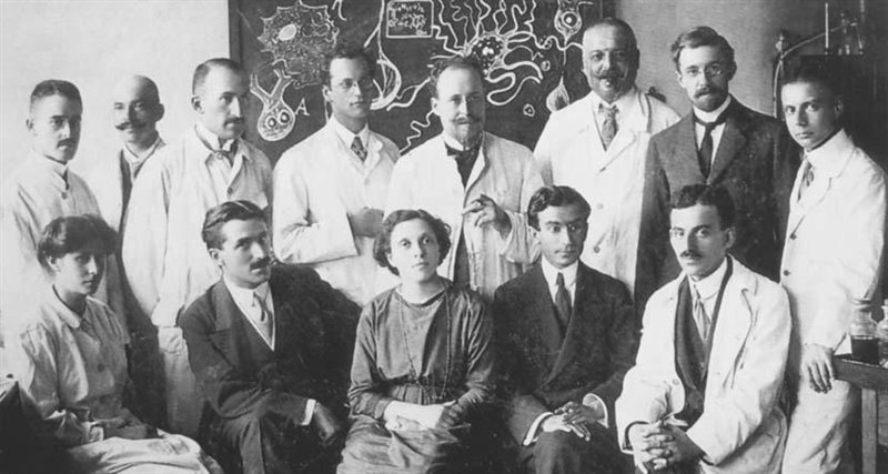 Grupo de investigadores en un laboratorio de neurociencia, posando para una foto en blanco y negro, con una pizarra de fondo que muestra esquemas neuronales.
