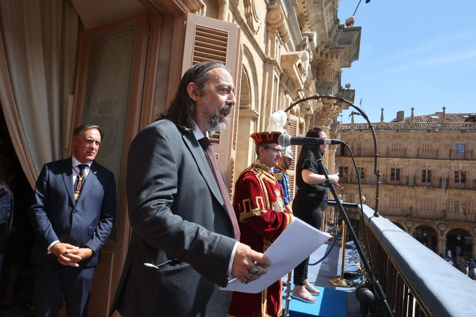 A ceremony in which a man with a beard and long hair, dressed in a suit, is seen holding papers while speaking from a balcony. Next to him is a woman with a microphone and a man dressed in a traditional costume. In the background, historic buildings can be seen.