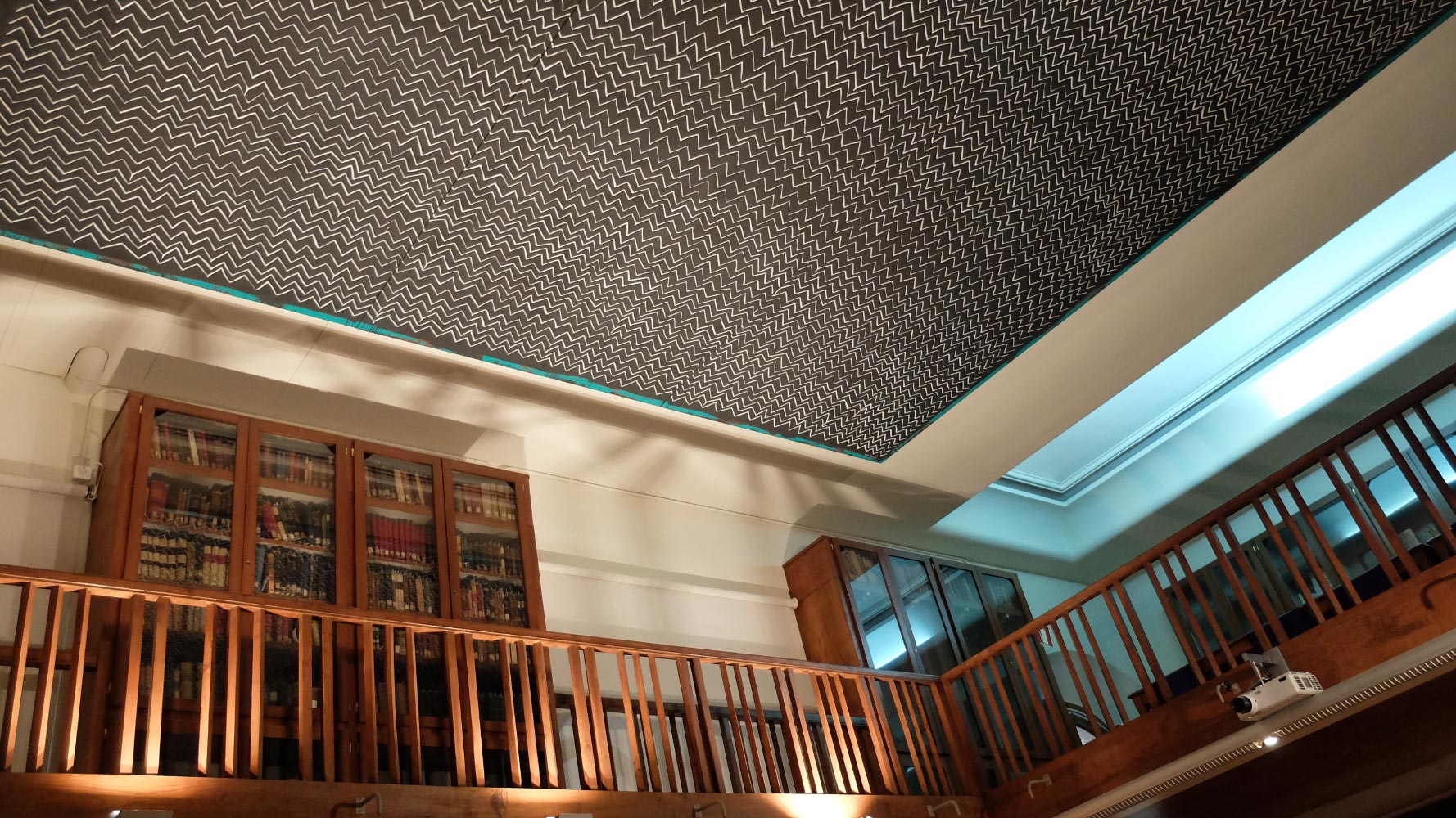 Vista del interior de una biblioteca, mostrando estanterías de madera con libros antiguos y un techo decorado con un diseño en zigzag en color negro y blanco.