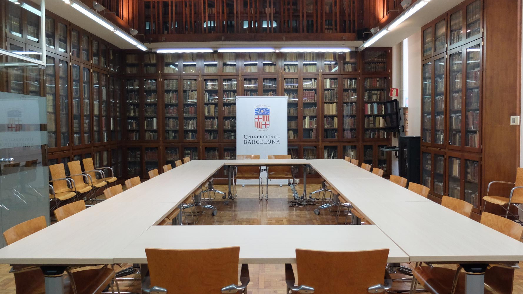 Interior de una sala de reuniones en la Universidad de Barcelona, con estanterías llenas de libros, mesas organizadas y sillas de madera.