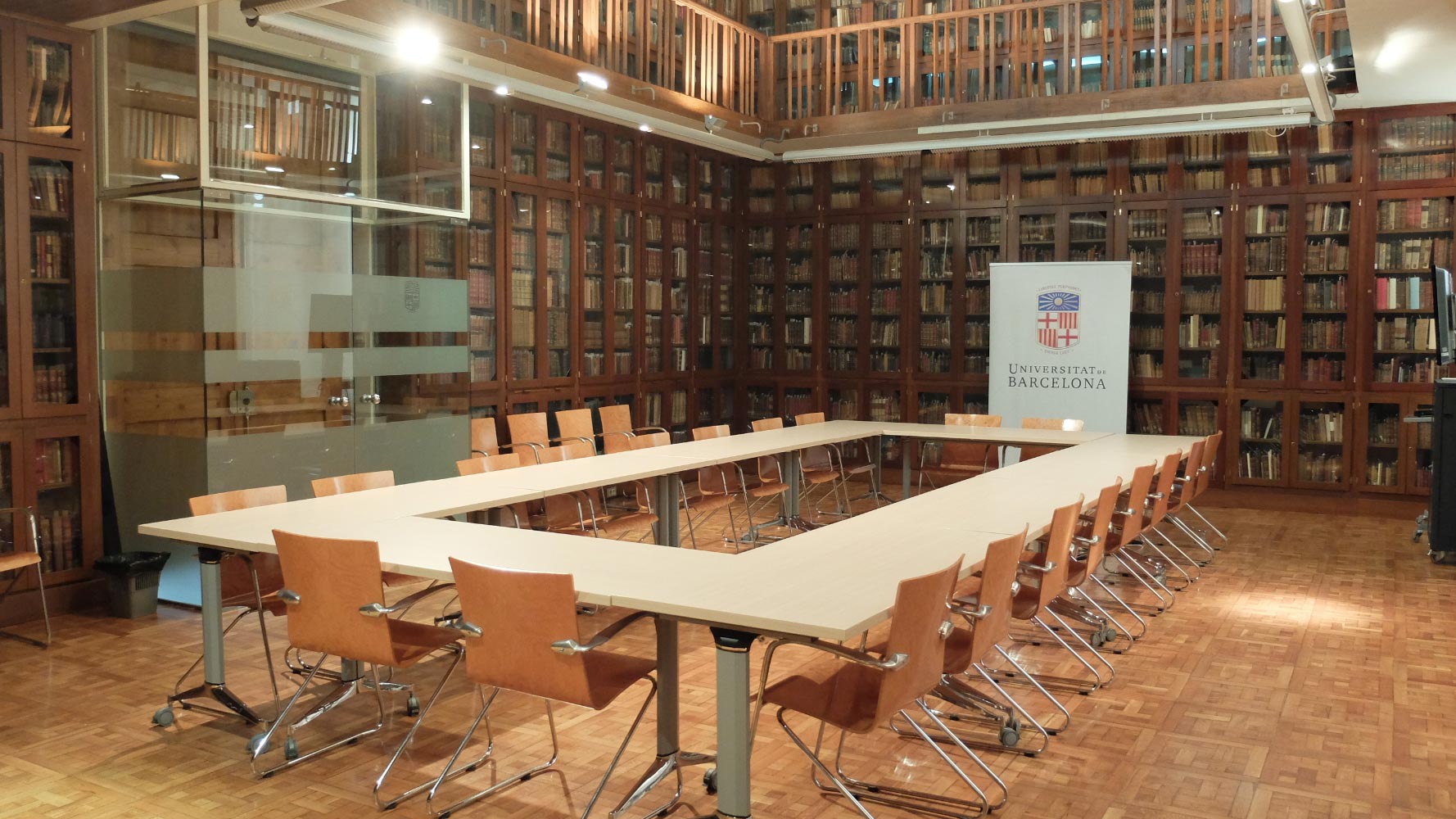 Interior de una sala de reuniones en la Universidad de Barcelona, con una mesa grande y sillas alrededor, rodeada de estanterías con libros y un banner de la universidad.