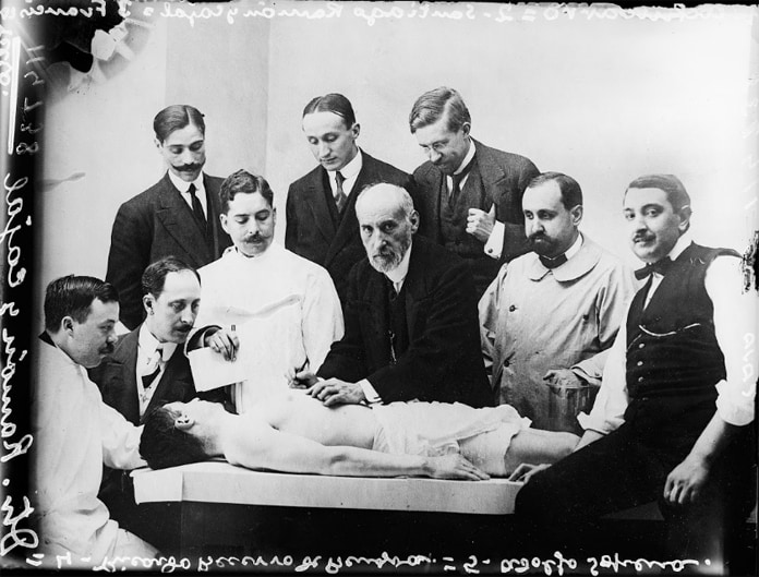 Group of physicians and medical students observing an anatomical exhibition in a laboratory, with a cadaver on a dissection table.