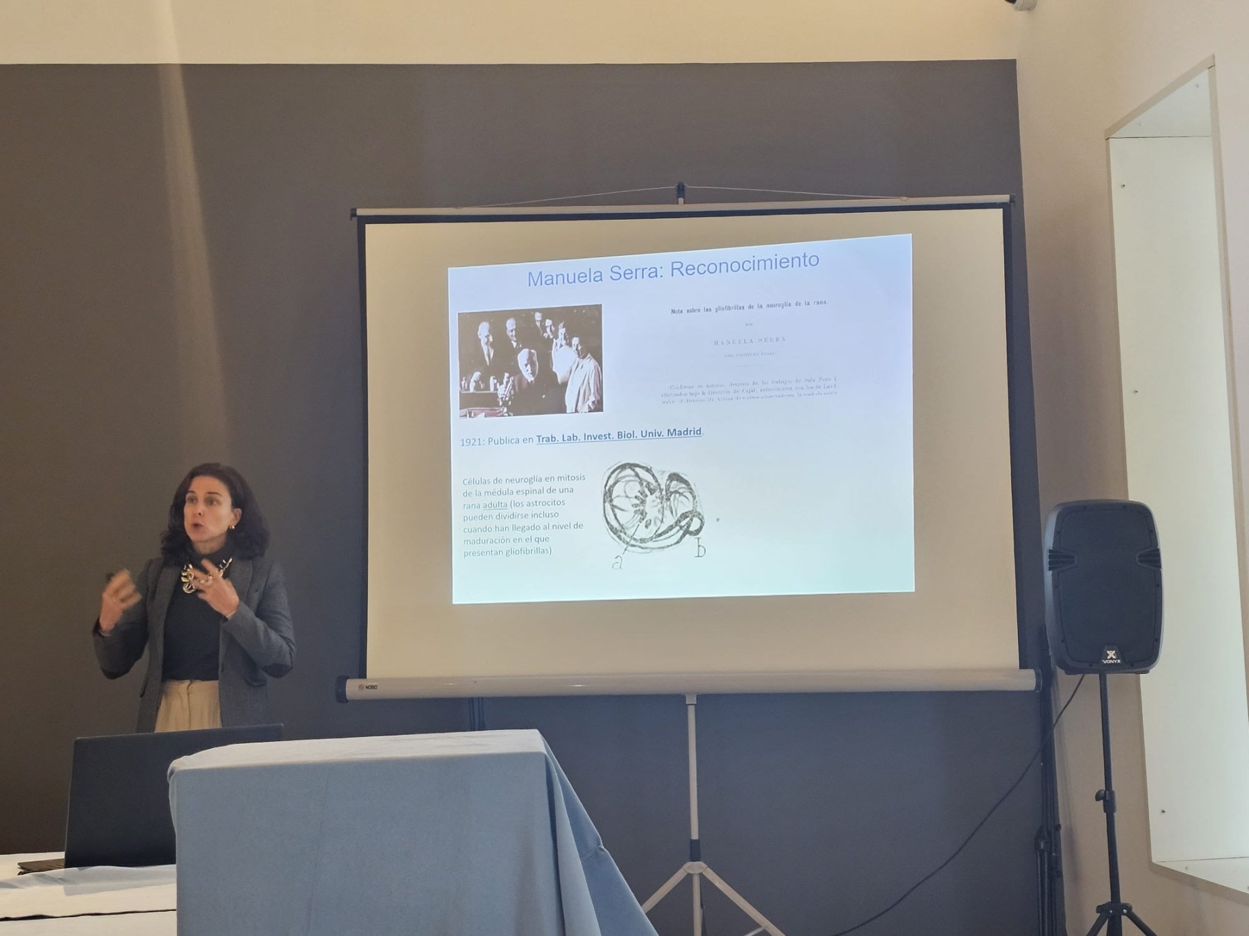 Woman presenting a slide about Manuela Serra at a science event, with a projector and table in the foreground.
