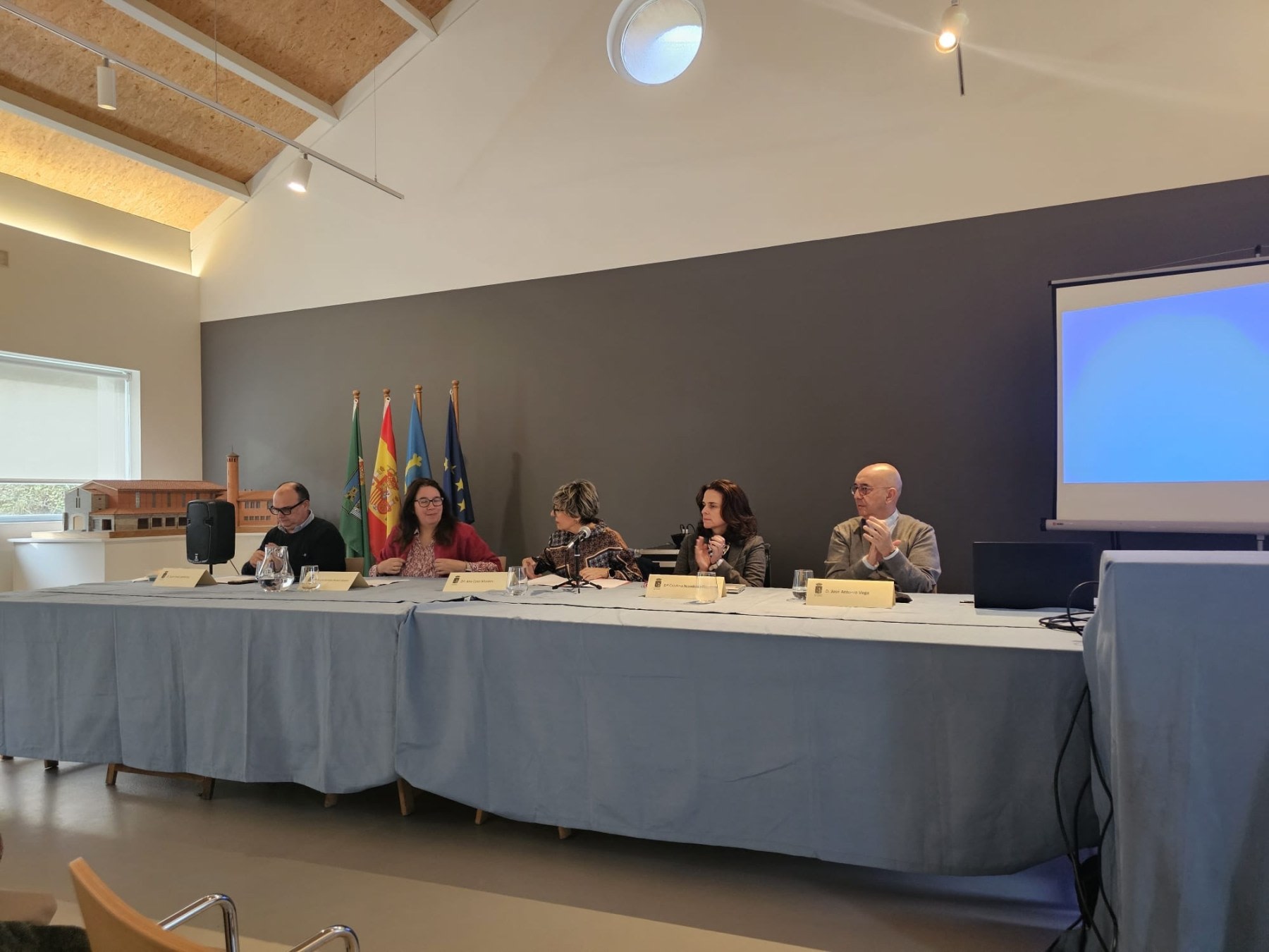 Panel of speakers at an event on science and the legacy of Santiago Ramón y Cajal, with flags in the background and a projected presentation.