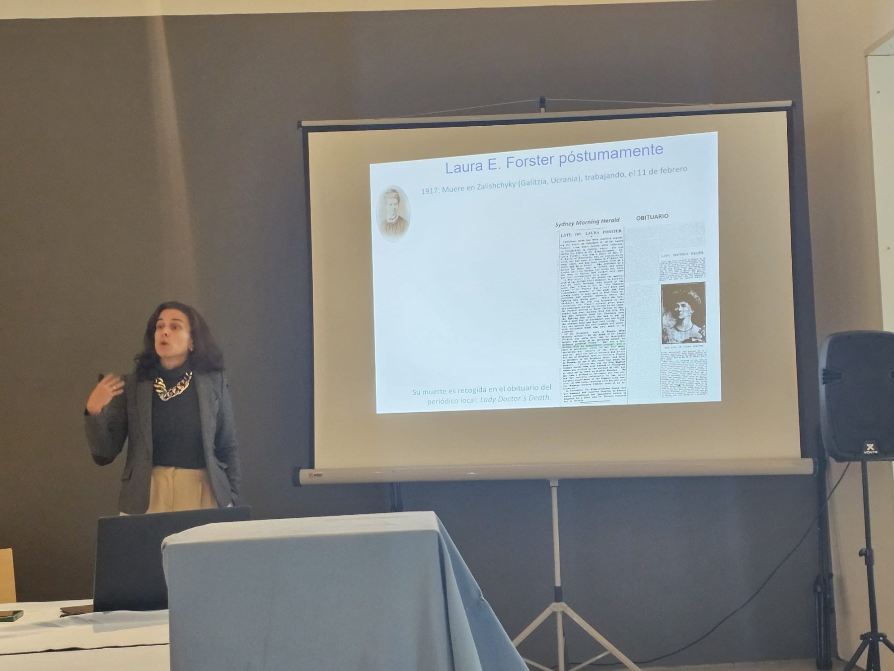 Speaker at a conference, talking about Laura E. Forster, with a screen presentation that includes details about her life and an obituary.