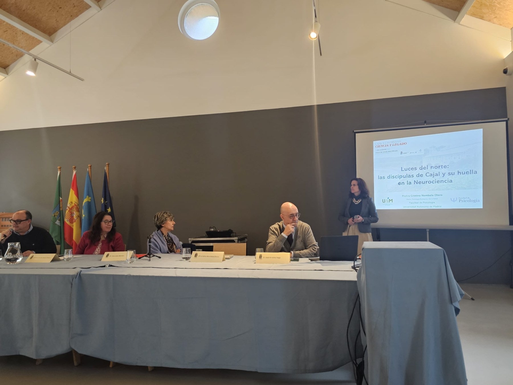 Speaker presenting at the conference "Lights from the North: Cajal's Female Disciples and Their Footprint in Neuroscience", with conference table and flags in the background.