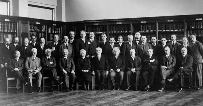 Group of scientists and academics gathered in a room with bookshelves, in a black-and-white photograph.