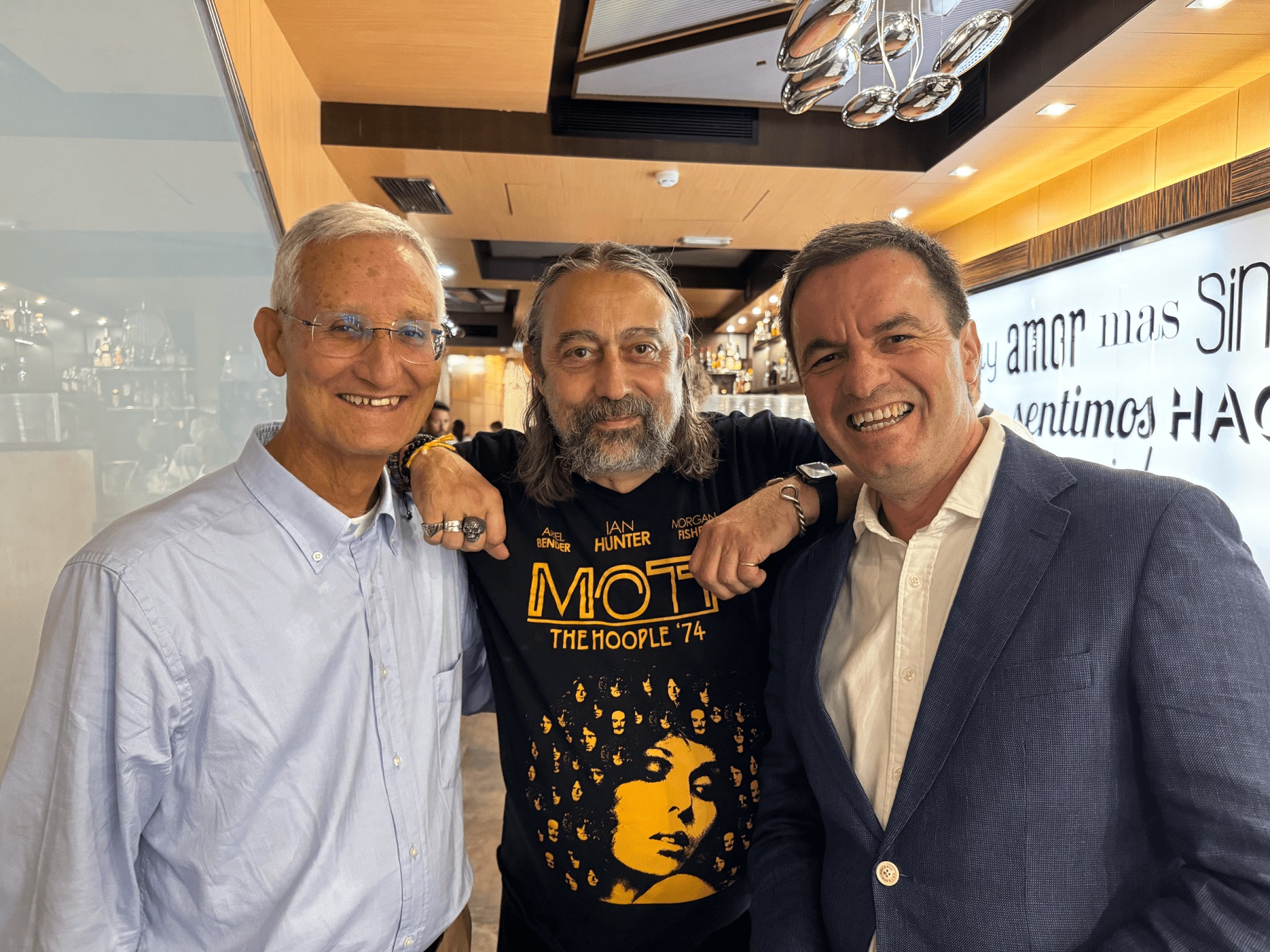 Three smiling men posing together in a restaurant, one wearing a black t-shirt with a yellow design, another in a white shirt, and the third in a blue jacket.