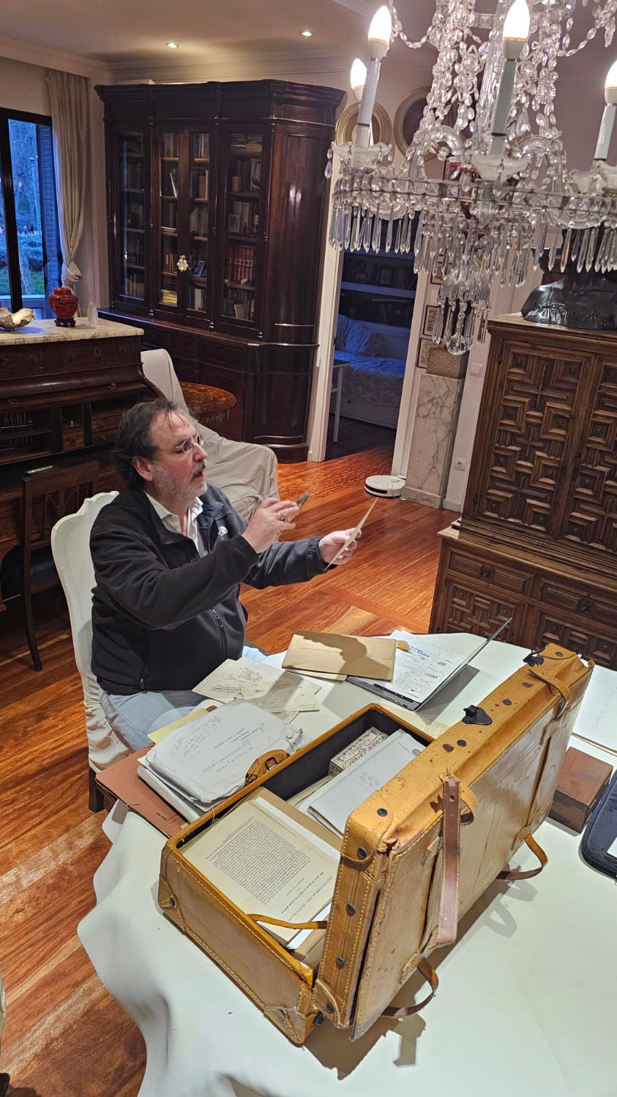 Man seated at a table, reviewing documents and holding a sheet, in an office environment with shelves and a candelabra.