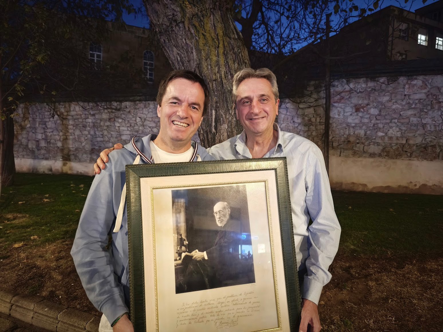 Manuel Garrosa García y José Adserias. Dos hombres sonrientes sosteniendo un marco con una foto en blanco y negro de un hombre mayor en un ambiente exterior al anochecer.