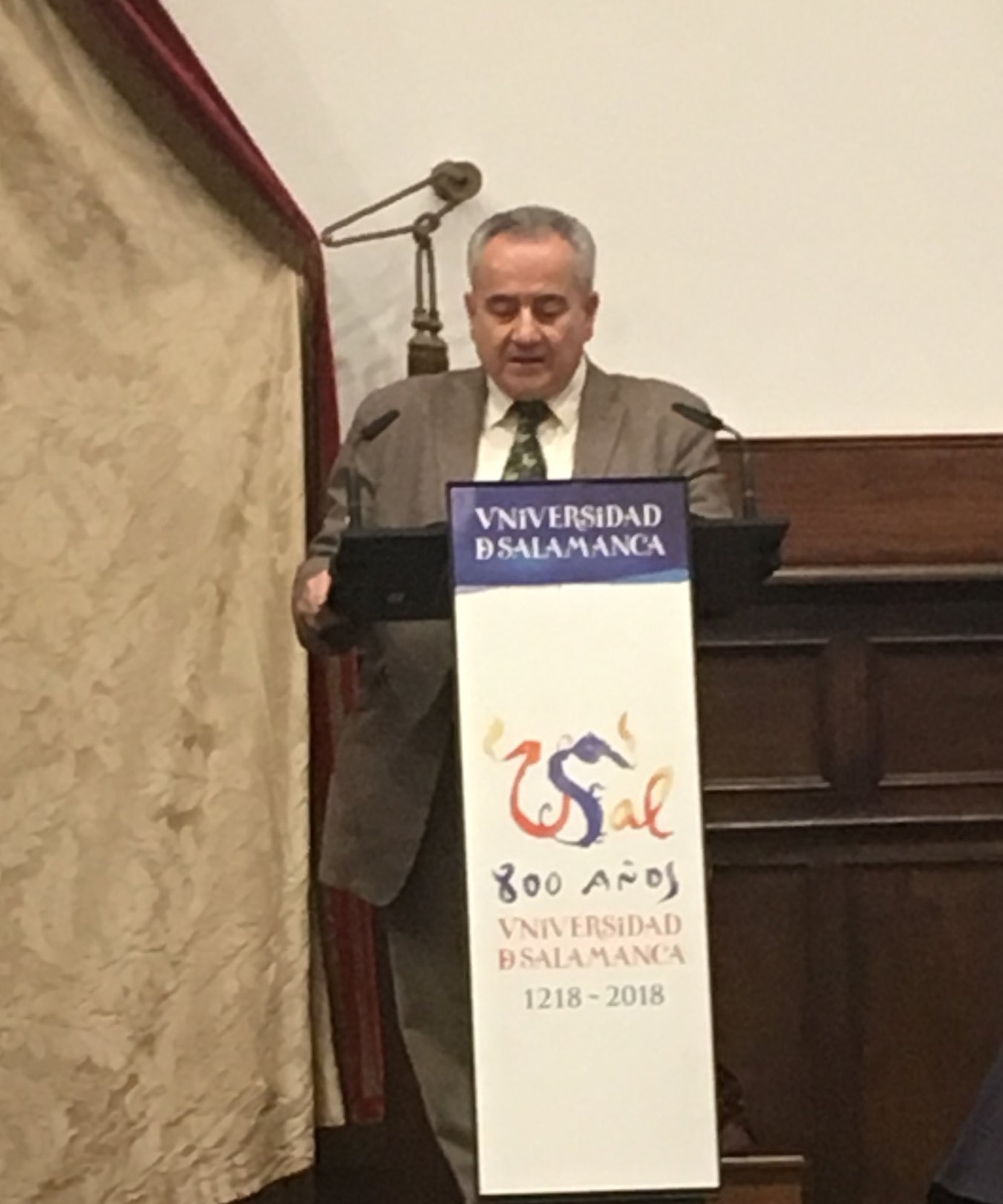 Person delivering a speech at the Paraninfo of the University of Salamanca, with a poster celebrating the institution's 800 years, in the context of an academic event.