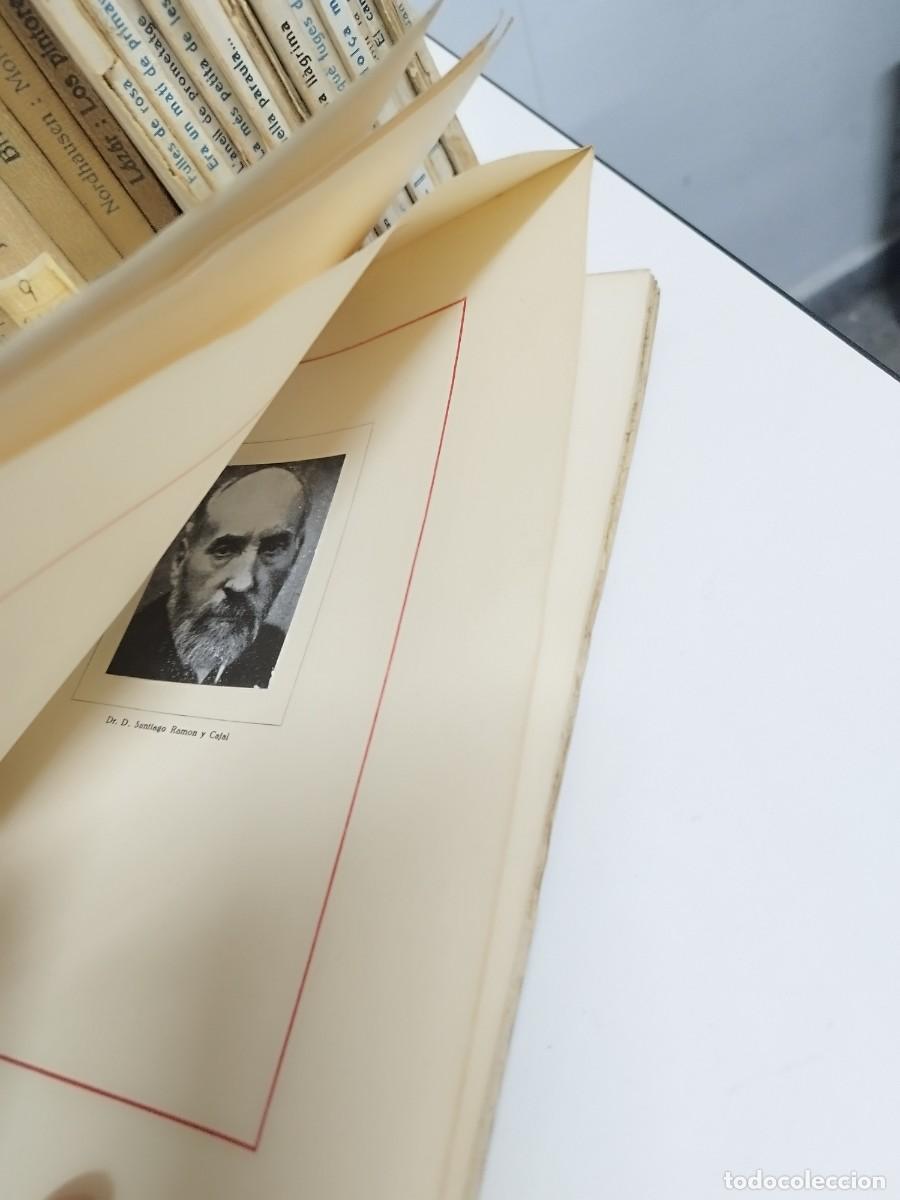 Un libro antiguo abierto que muestra una imagen de Santiago Ramón y Cajal en su interior, con páginas amarillentas y en el fondo, otros libros visibles.