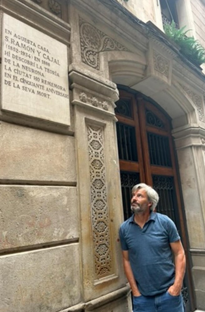 Hombre frente a una fachada decorativa en Barcelona, observando una placa conmemorativa de Santiago Ramón y Cajal que menciona la teoría de la neurona.
