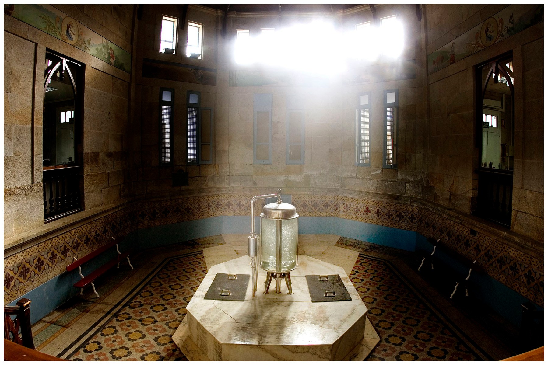 Interior del balneario de Aguas de Cabreiroá, mostrando una fuente octogonal de agua mineral con un diseño elegante, rodeada de paredes de piedra con ventanas y bancos de madera.