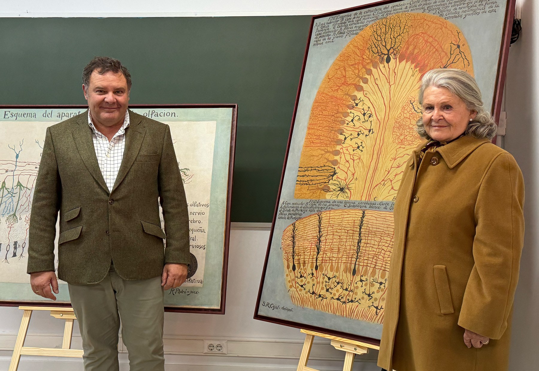 A man and a woman pose together in front of two works of art representing diagrams of the olfactory and neuronal system.