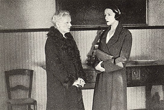 Dos mujeres de pie en un interior, una de ellas vestida con abrigo de piel y la otra con un vestido elegante, conversando en un ambiente académico.