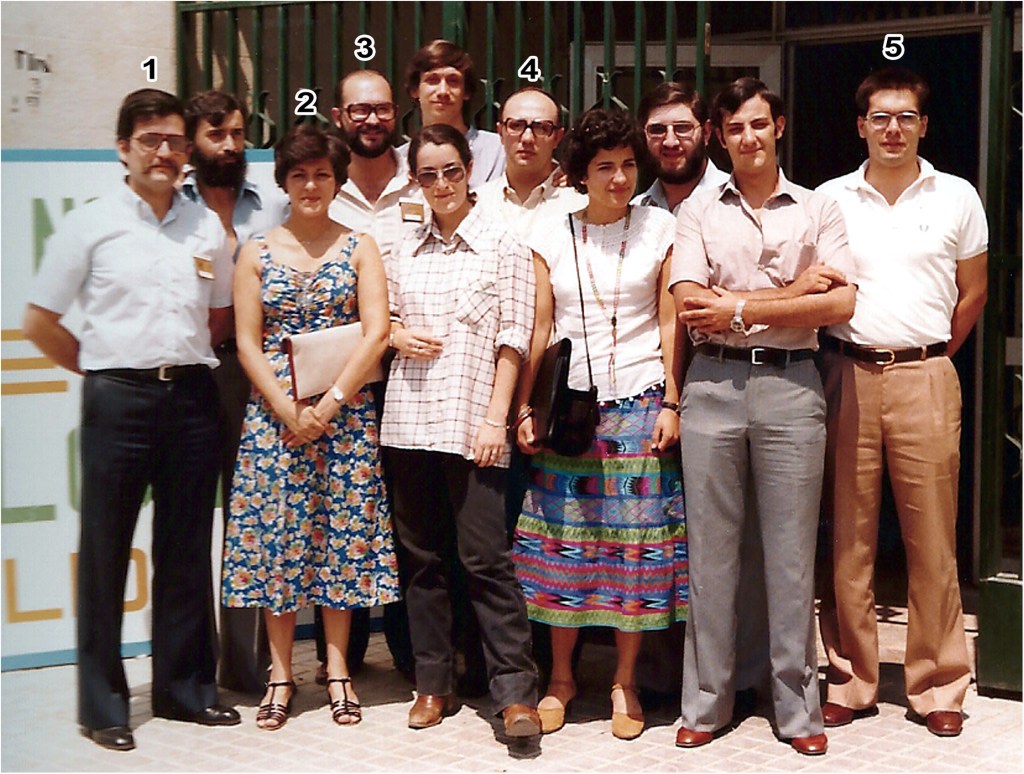 Group of people posing for a photograph, each in different styles of dress, in front of a green fence in an outdoor setting.