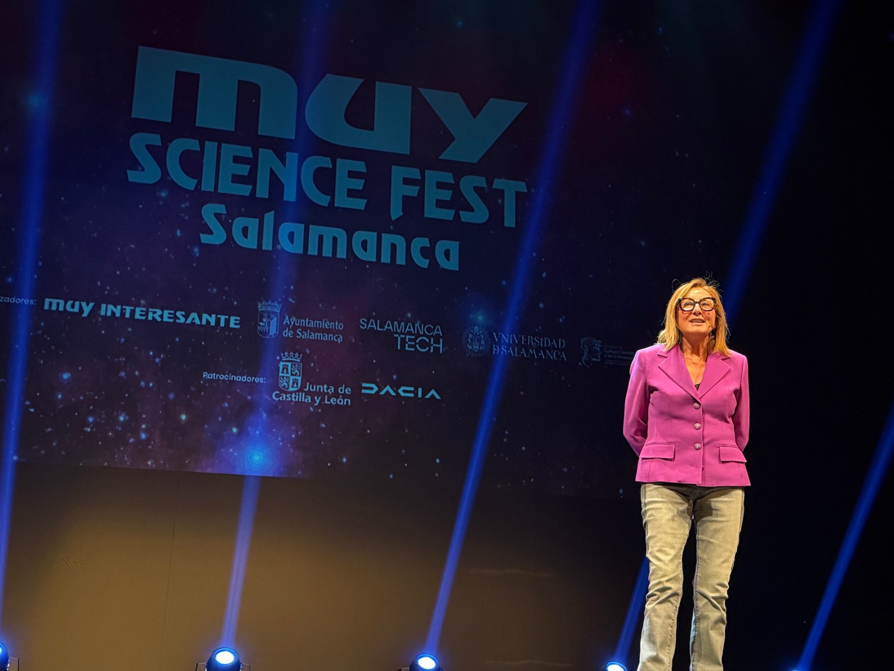 Presentadora en el escenario del MUY Science Fest Salamanca, con un fondo colorido que incluye el logotipo del evento y patrocinadores.