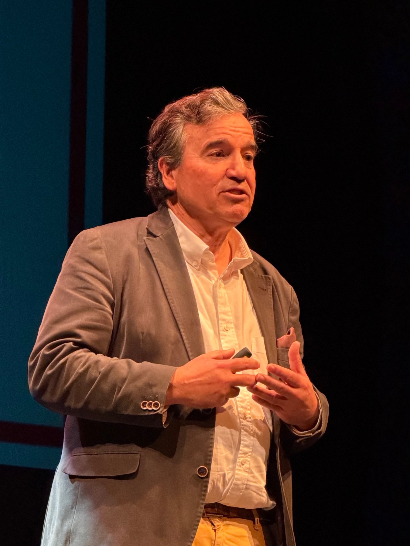 Un ponente hablando en el escenario del Teatro Liceo, vestido con chaqueta gris y camisa blanca, gesticulando mientras se expresa sobre temas científicos.