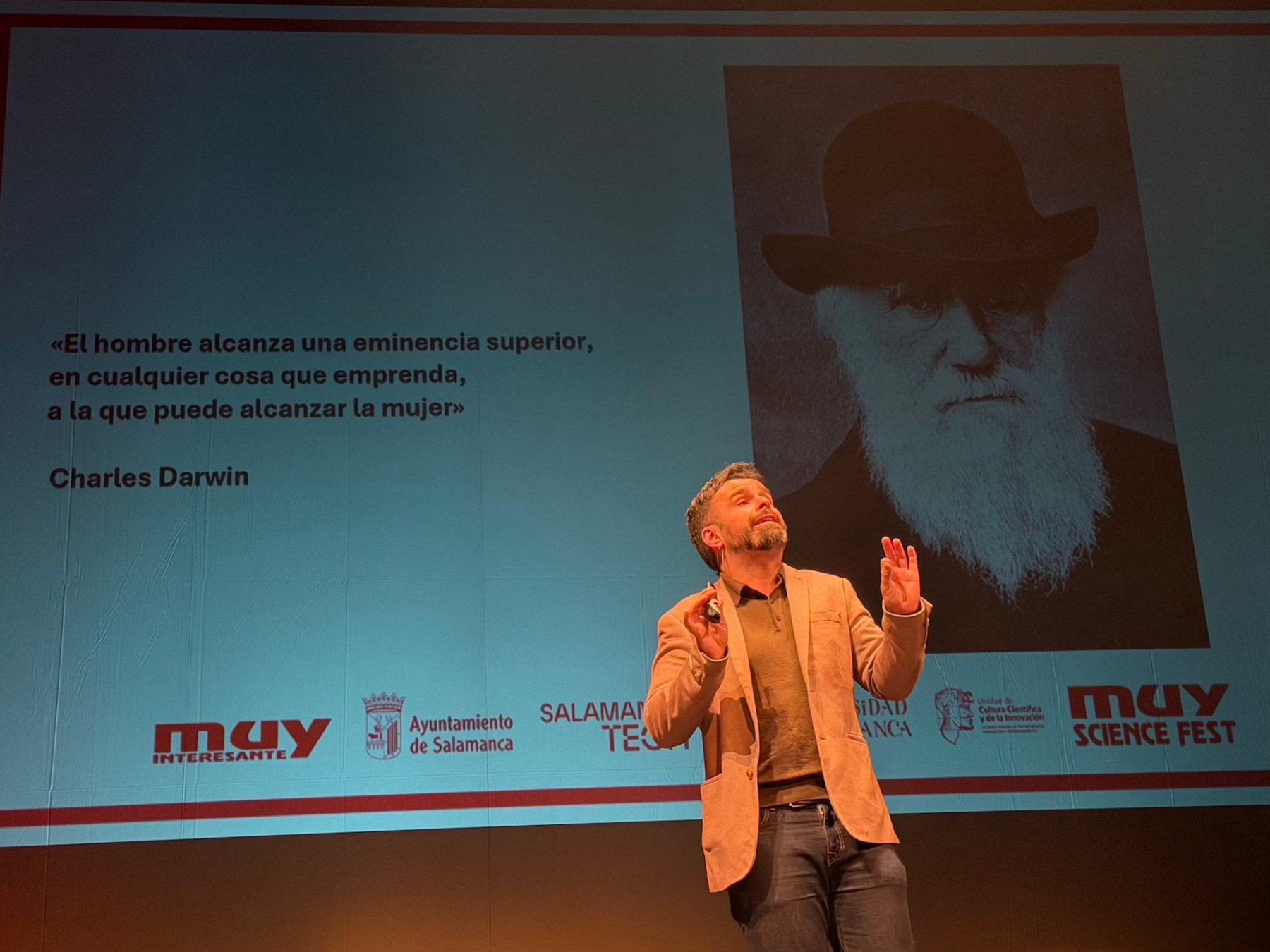 Presentador en el escenario del MUY Science Fest, mientras se proyecta una cita de Charles Darwin sobre la igualdad de género.