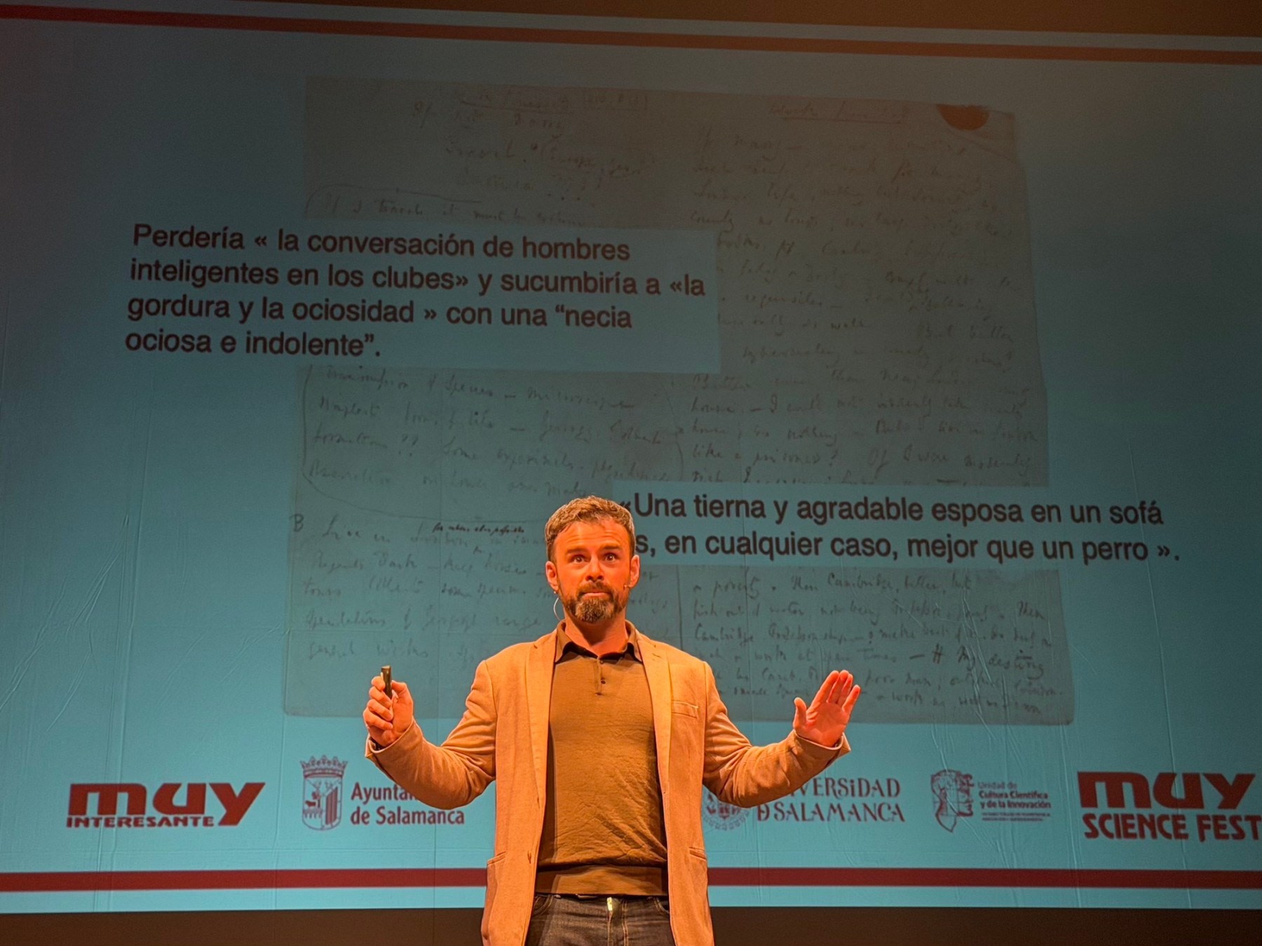 Un presentador en el escenario del MUY Science Fest 2025 muestra una diapositiva con un texto manuscrito, mientras sostiene un micrófono. El fondo incluye los logotipos de la revista 