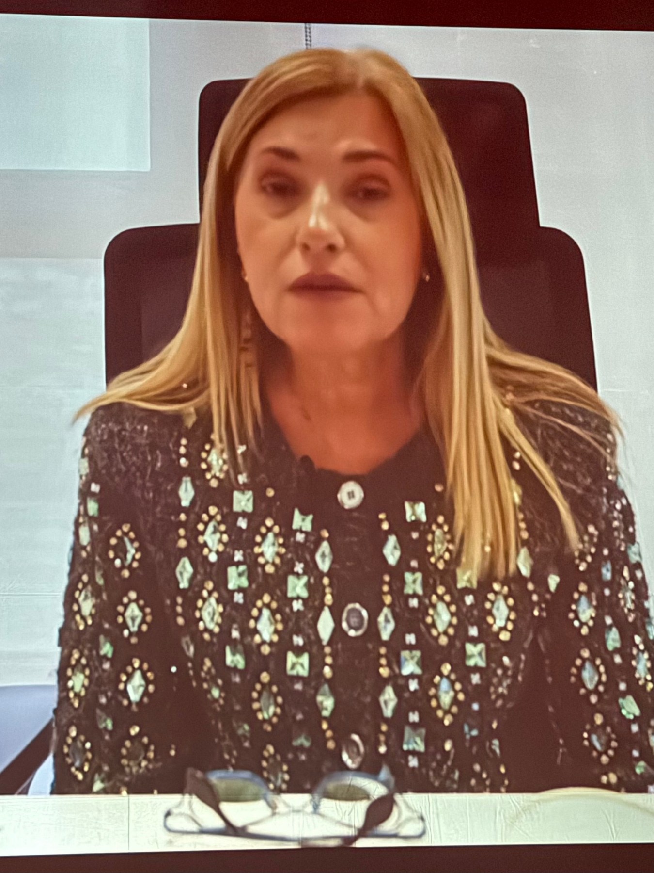 Una mujer con cabello largo y rubio, vestida con una chaqueta decorativa y un fondo gris, habla frente a una cámara.