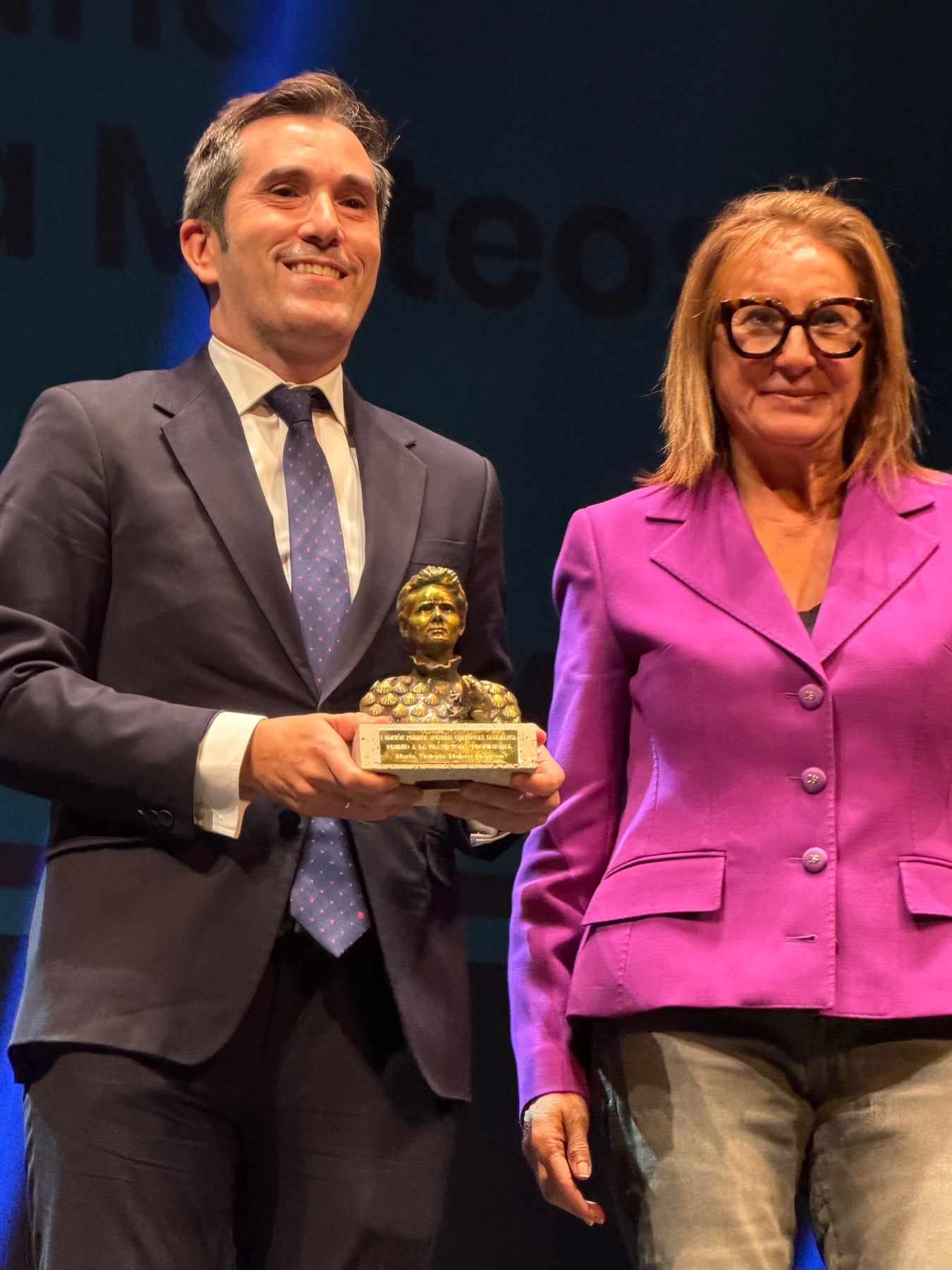 Dos personas están en un escenario durante un evento. Un hombre lleva un traje oscuro y sostiene un premio en forma de figura dorada, mientras que una mujer a su lado viste una chaqueta rosa y sonríe. Al fondo se puede distinguir un panel con texto en letras grandes.