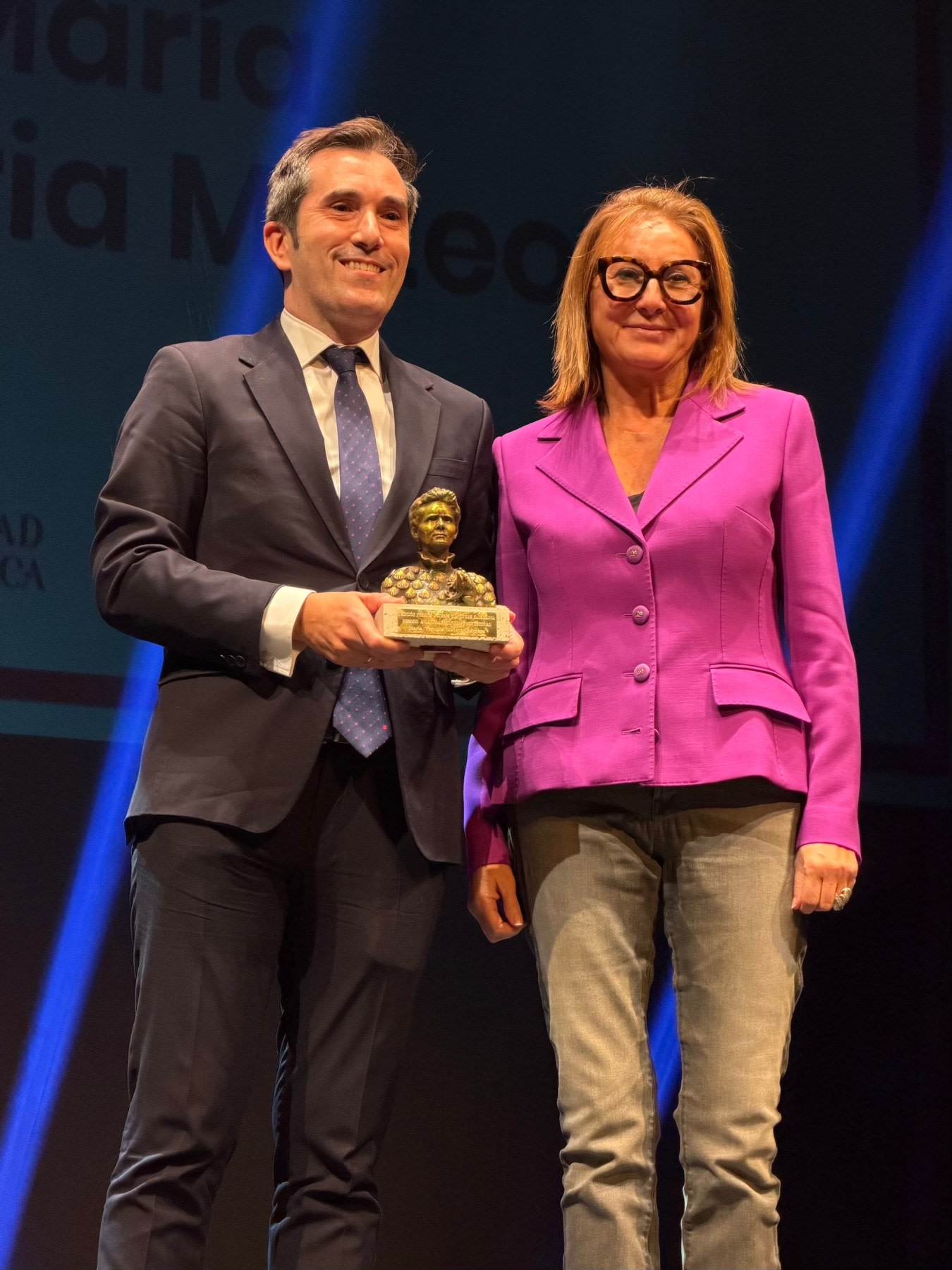 Un hombre y una mujer en un escenario sosteniendo un premio, con el fondo iluminado y el nombre de María Victoria Mateos visible en la pantalla.