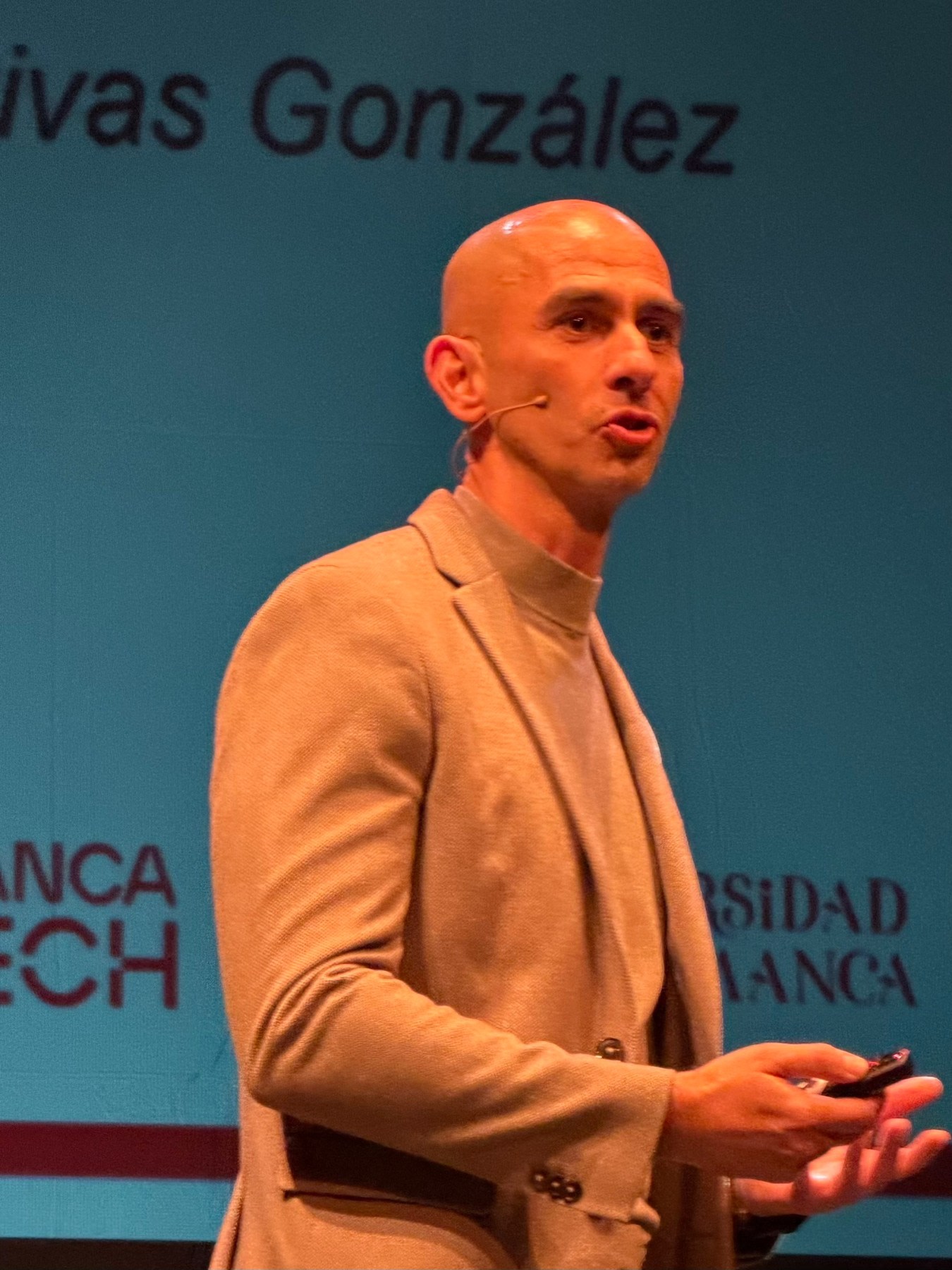 Un presentador con cabello rapado, vistiendo un suéter gris y chaqueta, habla en un escenario con un fondo que menciona 