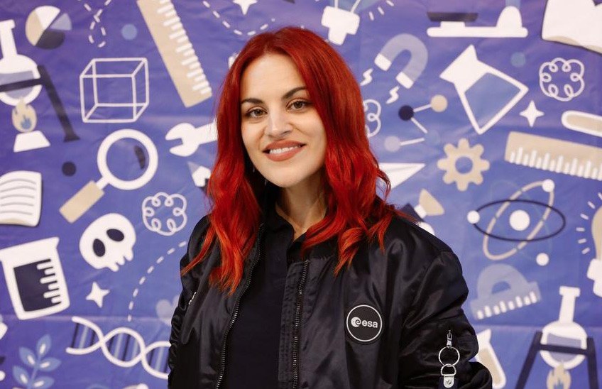 Retrato de una mujer con cabello rojo, vestida con una chaqueta negra y una camiseta oscura, sonriendo frente a un fondo ilustrado con símbolos científicos.