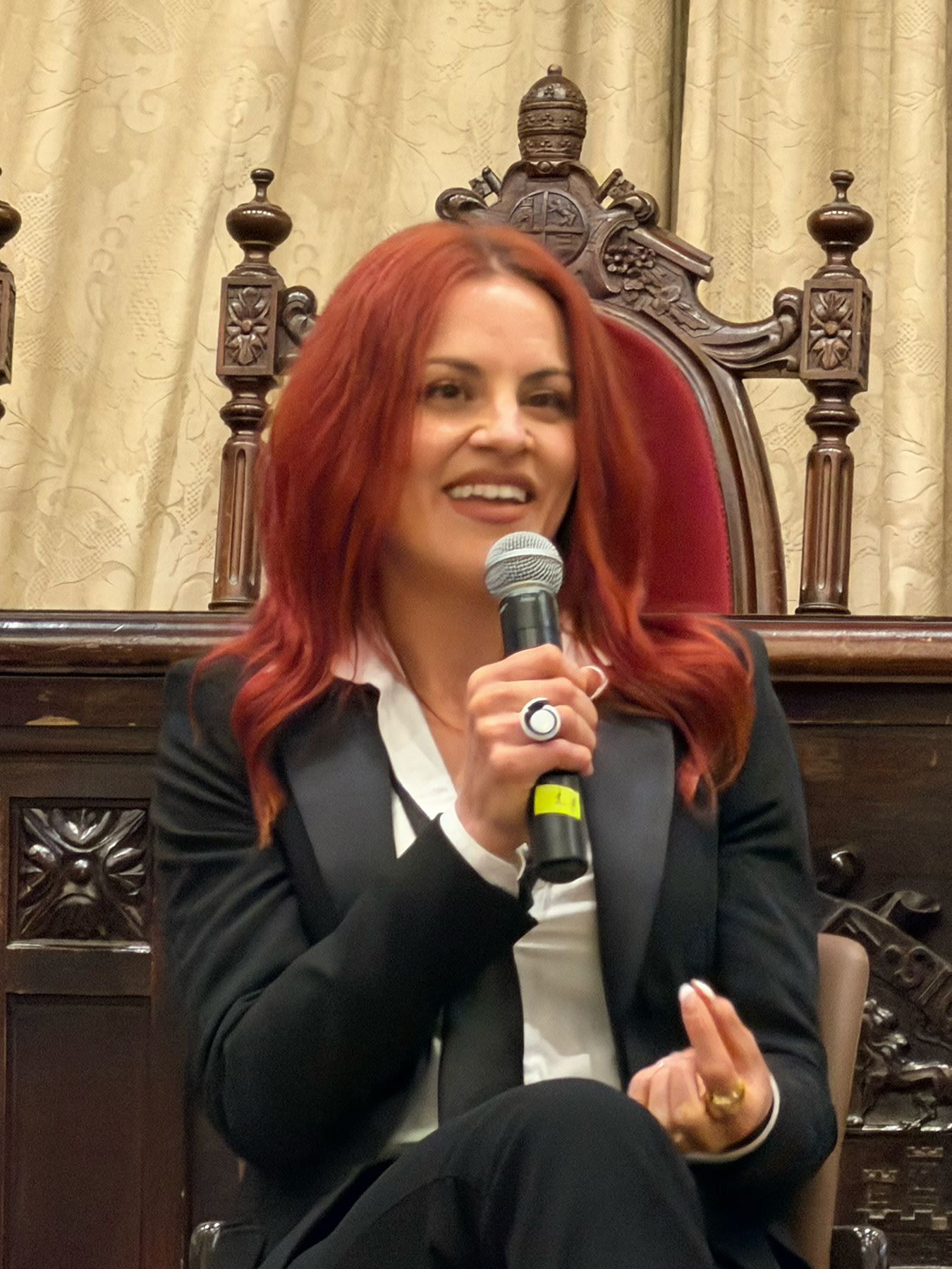 Mujer con cabello largo y rojo, vistiendo un traje negro y una blusa blanca, hablando en un micrófono en un evento, con un fondo de cortinas y sillas decorativas.