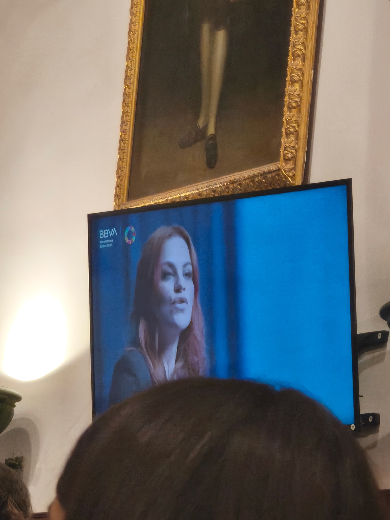 Una persona con cabello largo y rojo habla en un televisor en un ambiente decorado con un marco dorado que sostiene un retrato en la pared.