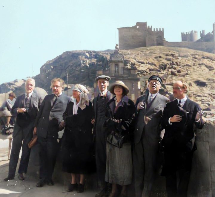 Group of people posing at an outdoor location, with a hill and a fortification visible in the background. All members of the group wear formal attire and some wear hats. The scene appears to capture a moment of joy or celebration.