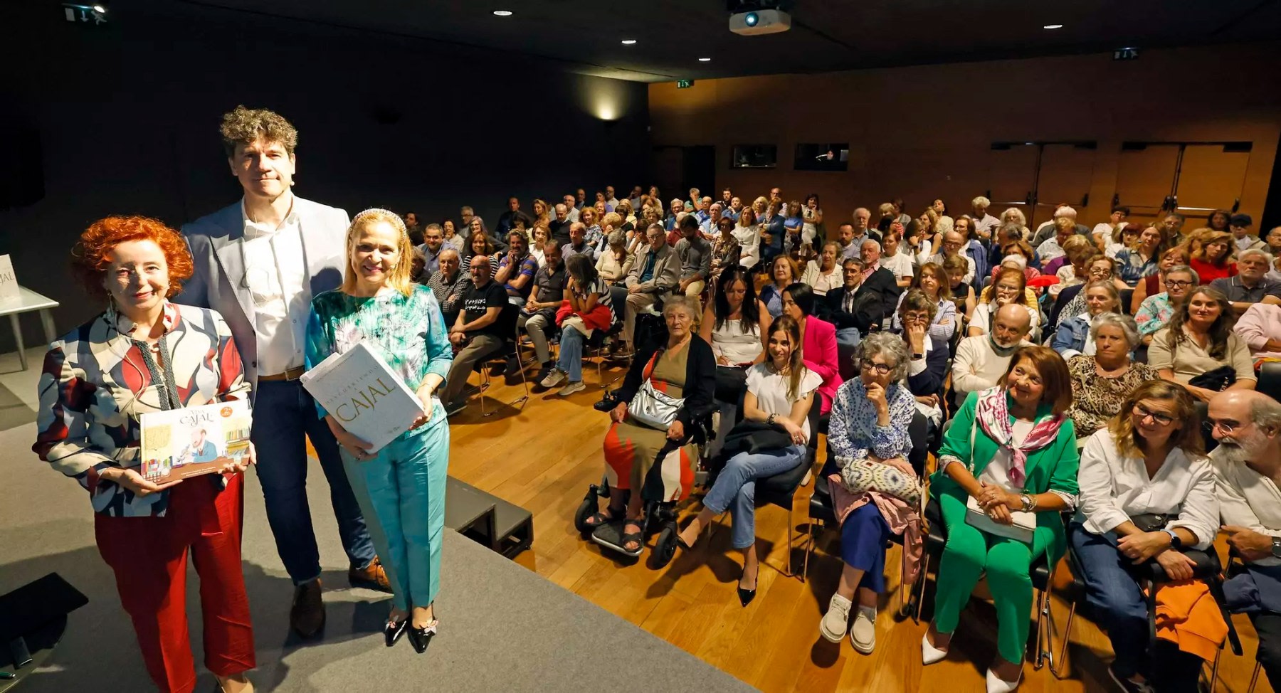 Tres personas posando en el escenario sosteniendo libros sobre Santiago Ramón y Cajal, con un público numeroso visiblemente atento al fondo en un evento cultural.